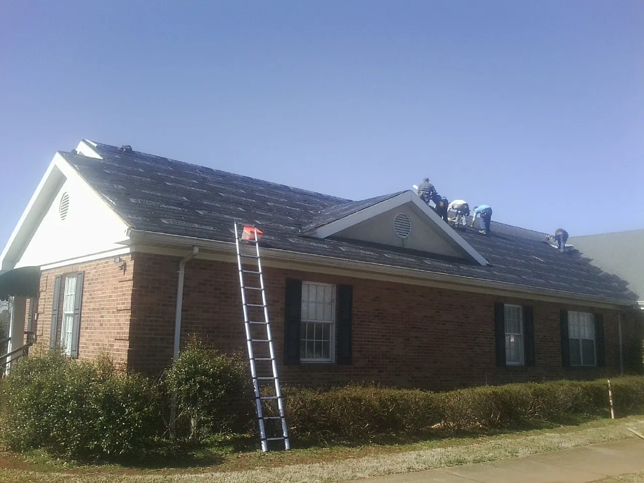 Crew tearing shingles off a brick ranch roof with a ladder set — Metal Roof Repair work by our crew in Webster City