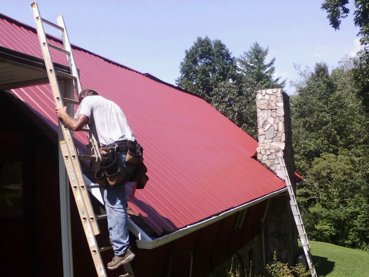 Roofer on a ladder beside a red metal roof and stone chimney — Soffit & Fascia Repair by Roofing Brothers in Webster City